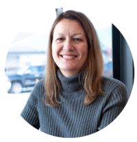 Photo of Trudy in a circle, by a window. She is wearing a grey turtleneck, smiling.