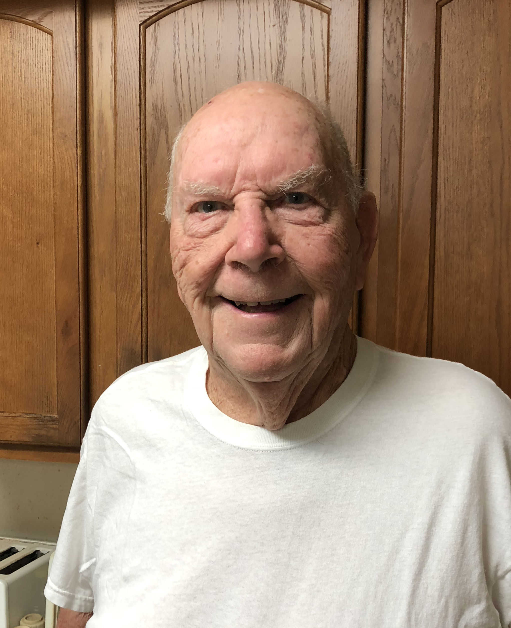 A white man stands in front of kitchen cabinets. He is wearing a white t-shirt, and is smiling.