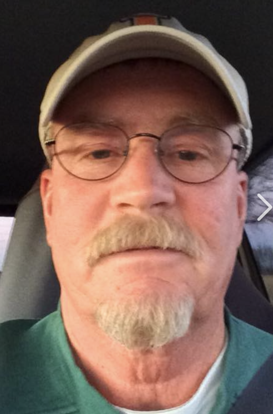 A white man is in his car, looking at the camera. He has a blonde/grayish goatee, glasses, and a baseball cap on.